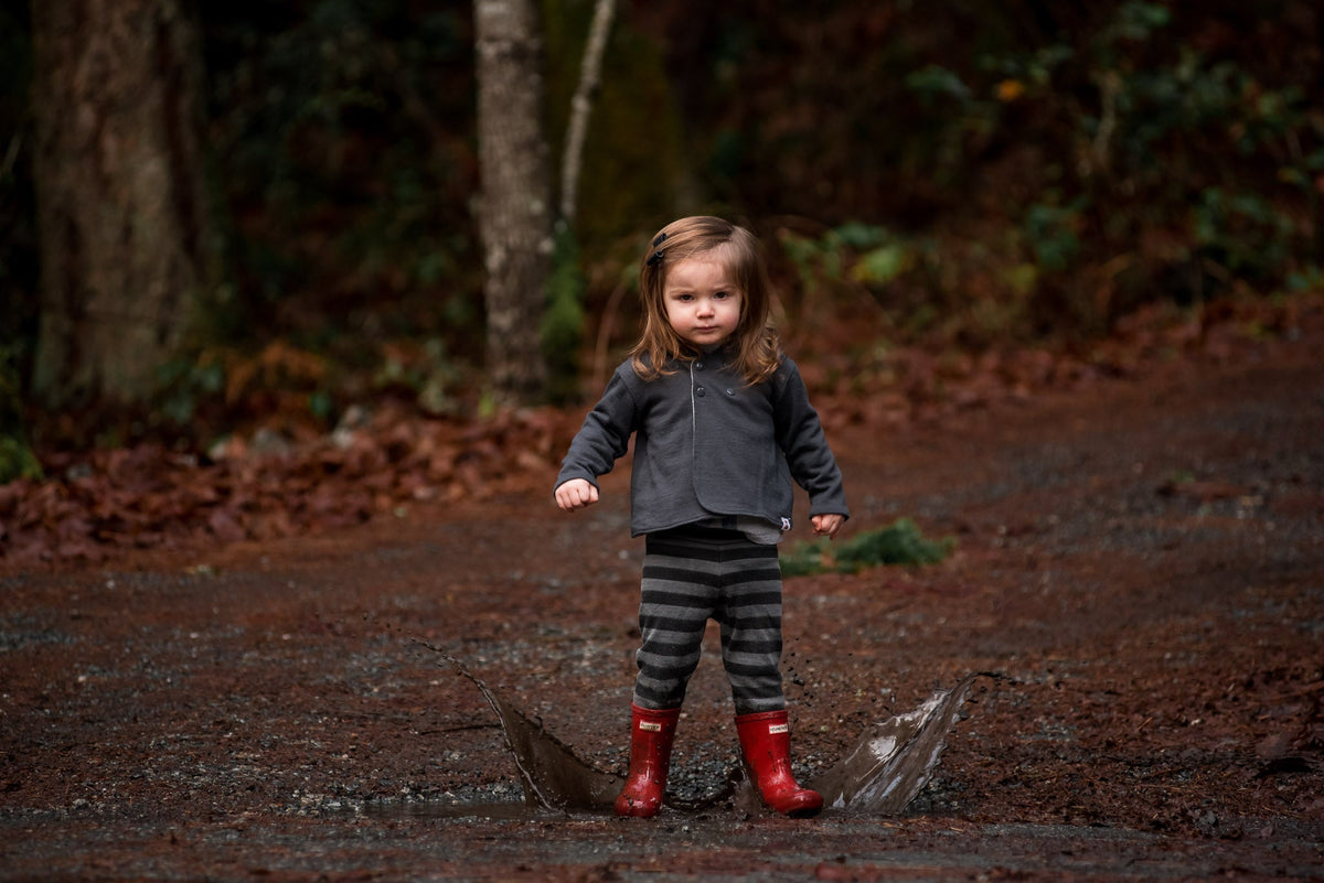 Toddler in a muddy forest wearing a merino jacket and merino leggings. She is jumping in a mud puddle, and the mud is splashing either side.