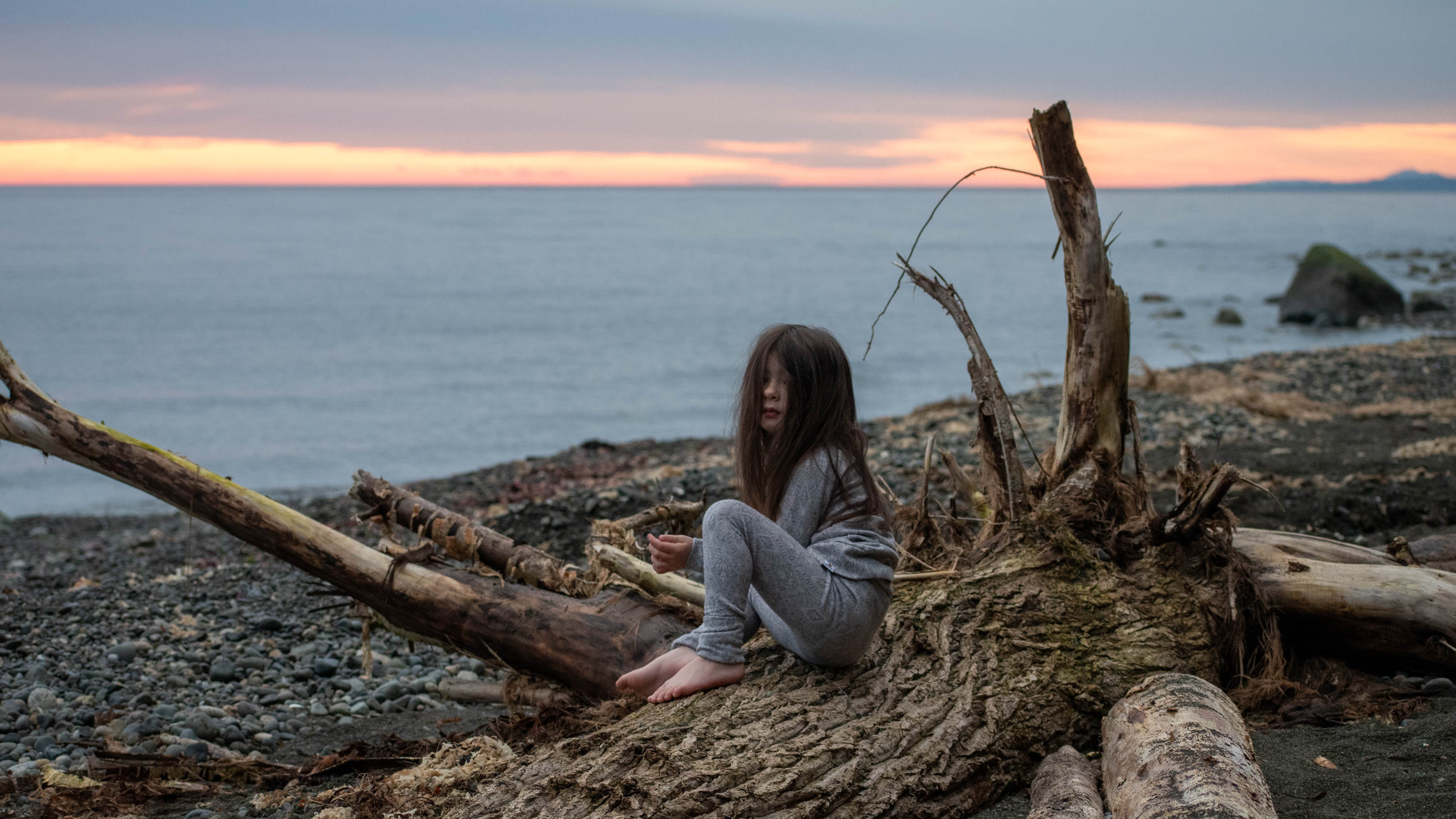 Girl sitting on driftwood at sunrise wearing a cozy mid-heavy weight merino wool base layer set. TK Clothing