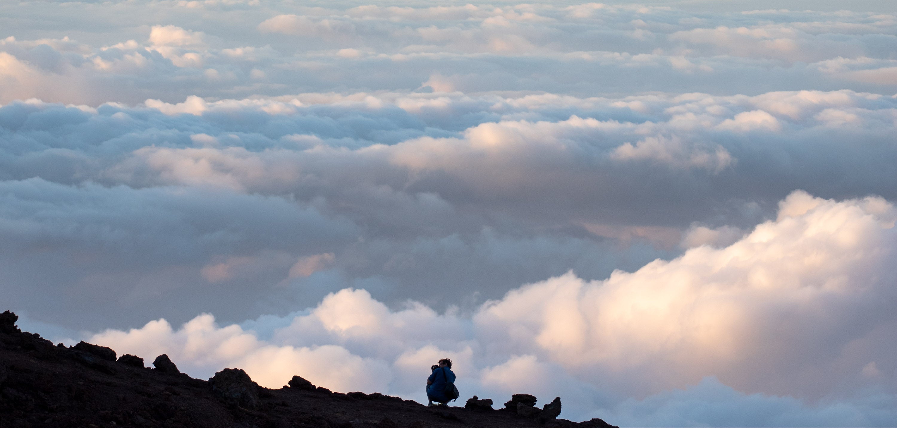 Adult hiker crouching on a high mountain ridge above the clouds at sunset wearing a blue merino wool base layer top. TK Clothing