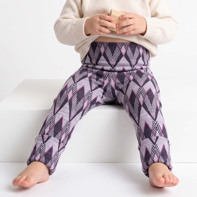 A toddler sitting on a white block wearing a cream knit sweater and purple geometric print merino  leggings, holding a small wooden block against a clean white background.