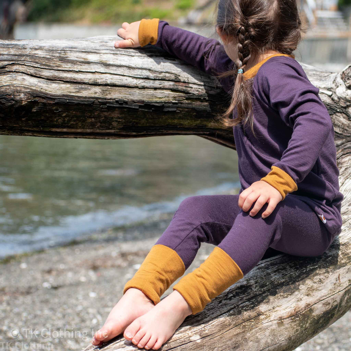 Child in iris and ochre handmade outfit sitting on driftwood by the water, TK Clothing Inc
