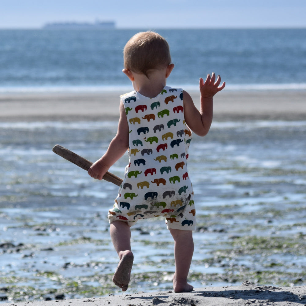 Toddler walking away on a sandy beach holding a piece of driftwood, wearing a sleeveless organic cotton romper with a colorful elephant print made by TK Clothing
